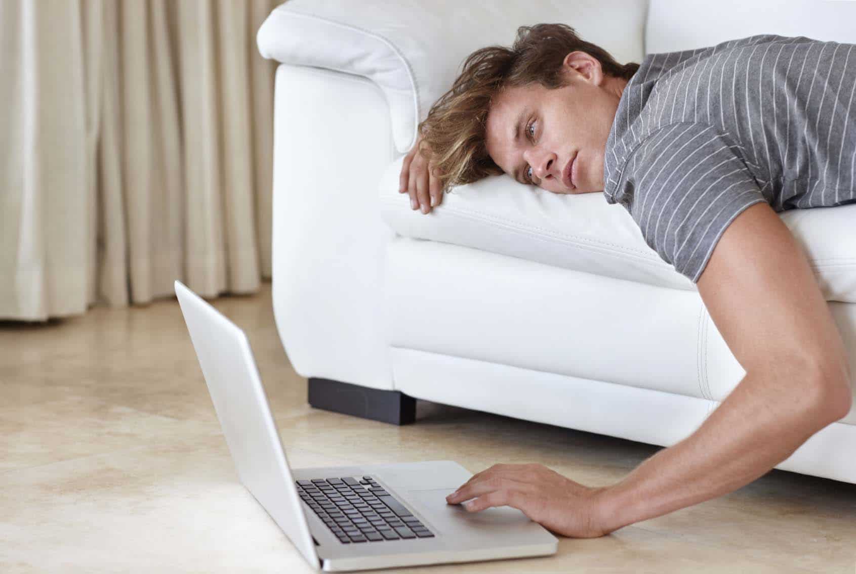 A young man lying on his couch with his hand resting on his laptop's keyboard