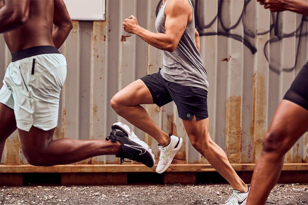 Three athletes running outside near a corrugated metal wall, focusing on their legs in motion.